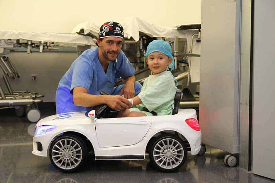 A surgeon in a hospital, crouching down next to a small child wearing hospital gown and cap, in a small electrical car that will be used to take him to the operating theatre