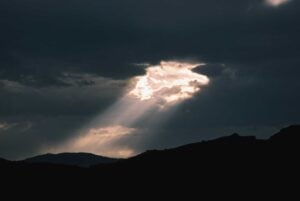 Sun rays breaking through dark clouds, above a hilly landscape.