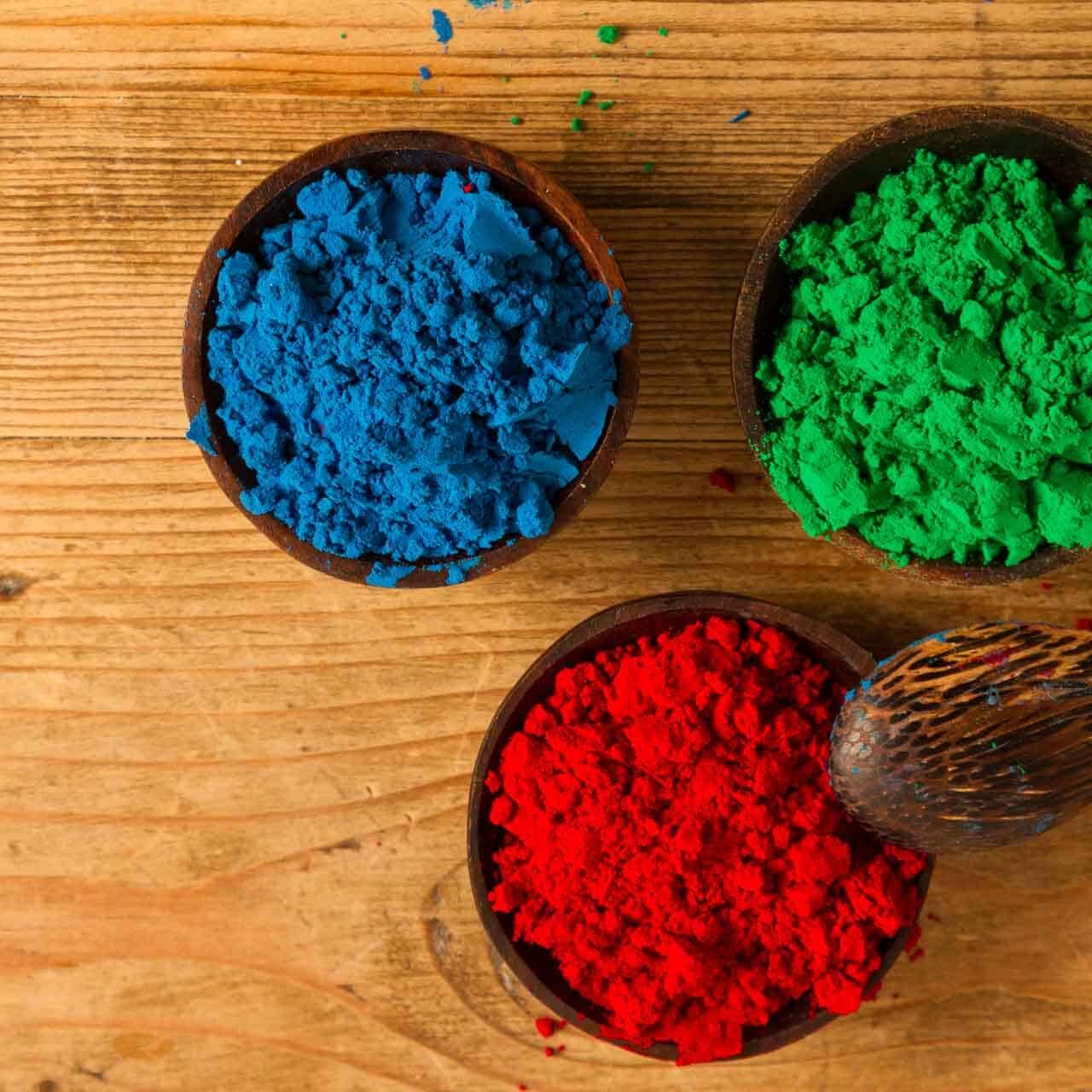 Red, green and blue powder pigments each in a small wooden bowl on a wooden surface.