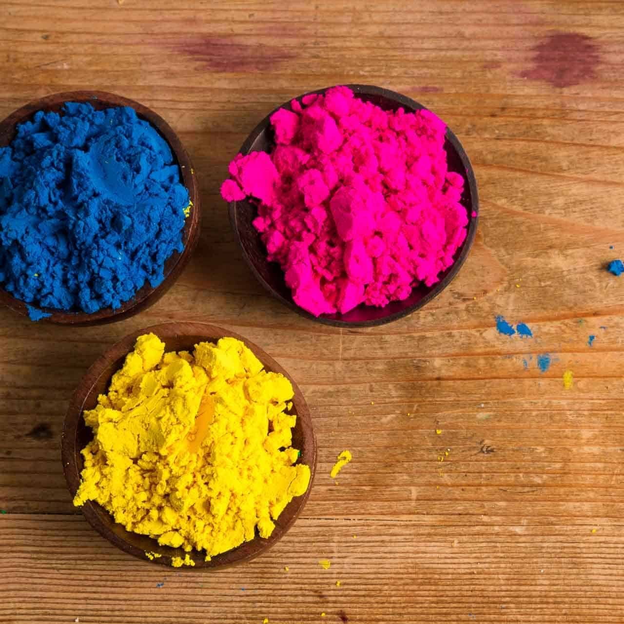 3 small wooden bowls on a wood surface, containing blue, magenta, and yellow powder pigments.